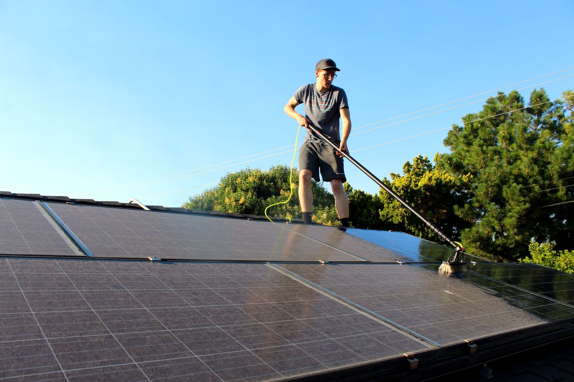 Bay Area Exterior Cleaning technician on a rooftop cleaning solar panels with a soft water-fed brush under a clear blue sky.